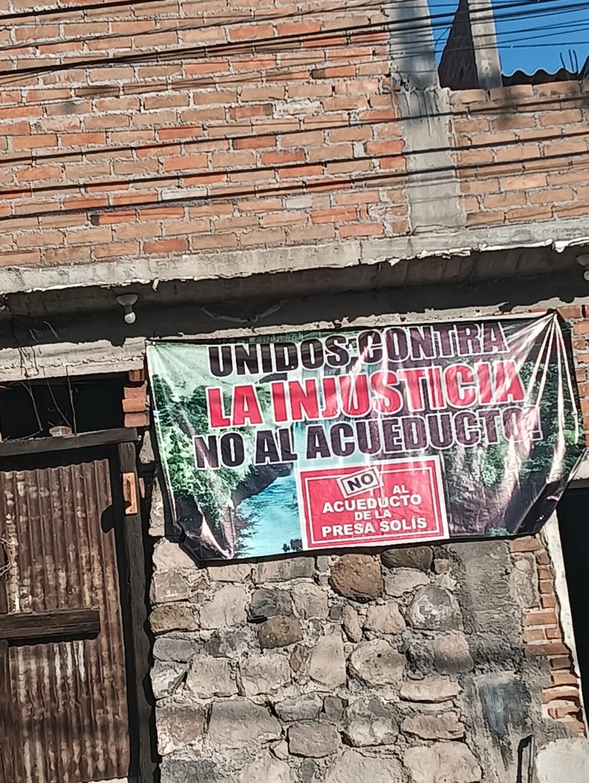 Manta colgada en un muro de tabique: «Unidos contra la injusticia. No al acueducto», con un cartel de «No al acueducto de la presa Solís».