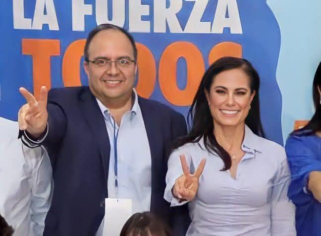Un hombre y una mujer hacen la señal de victoria frente a una lona de campaña.