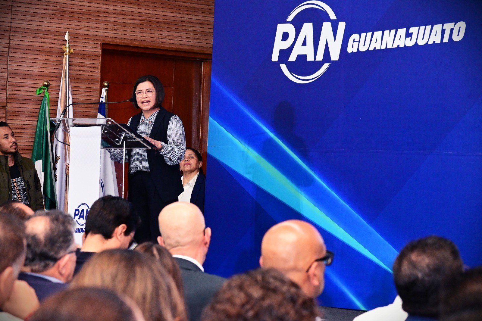 Una mujer habla desde el atril del PAN Guanajuato, con banderas a un lado y el público de espaldas en primer plano.