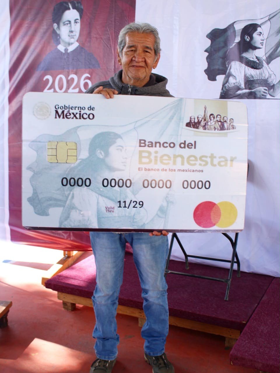 Un hombre mayor sostiene una tarjeta gigante del Banco del Bienestar en un evento del Gobierno de México.