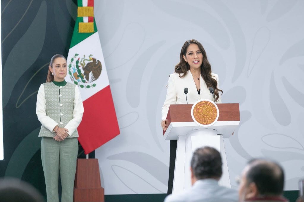 Libia Dennise García Muñoz Ledo habla en un atril con el escudo nacional mientras Claudia Sheinbaum Pardo la escucha de pie, junto a la bandera de México.