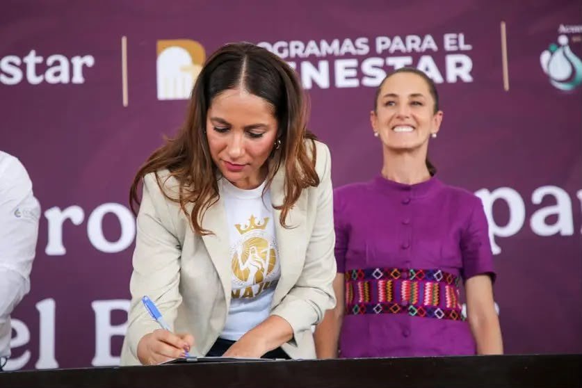 Libia Dennise García Muñoz Ledo firma un documento en un evento de Programas para el Bienestar; detrás, Claudia Sheinbaum Pardo sonríe.