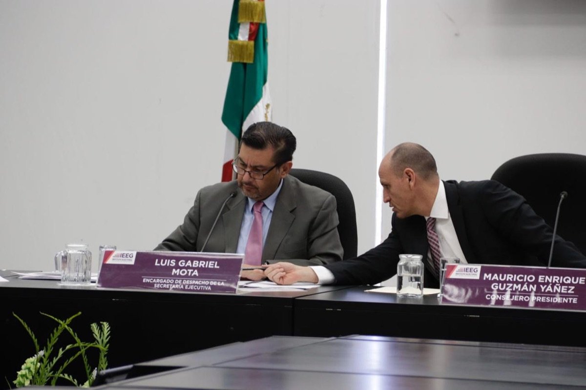 Dos funcionarios del IEEG conversan en la mesa del Consejo General junto a sus placas de identificación.