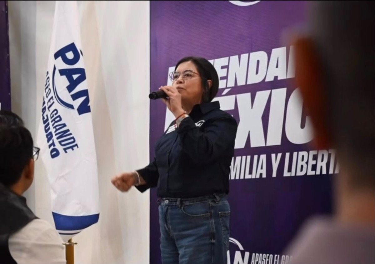 Juana de la Cruz Martínez Andrade habla con micrófono junto a una bandera del PAN.