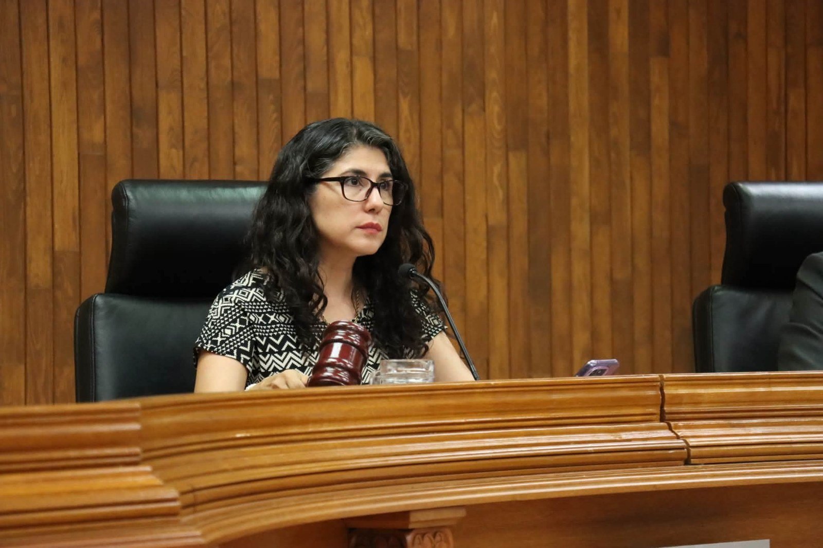 Una mujer de cabello oscuro y lentes, con blusa de patrón geométrico en blanco y negro, sentada tras el estrado de madera de un tribunal, con un mazo y un micrófono frente a ella.