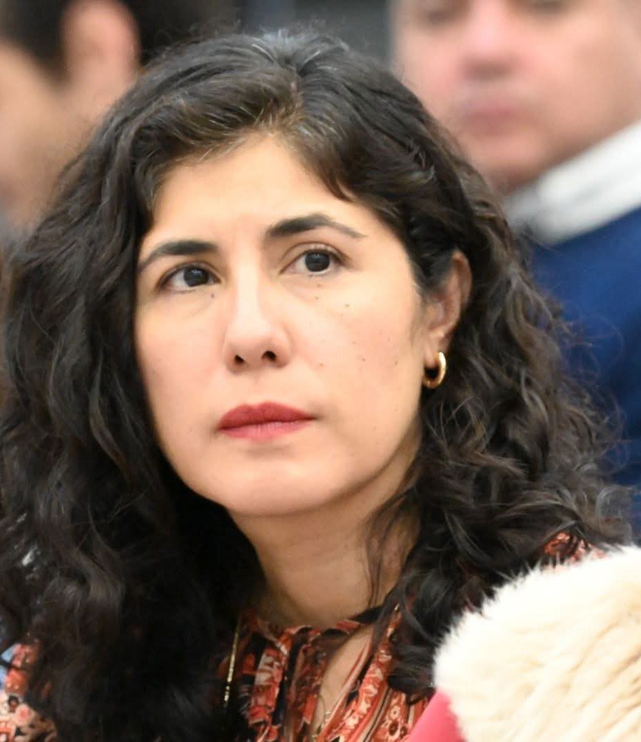 Primer plano de una mujer de cabello oscuro y ondulado, con aretes de aro dorados, mirando hacia un lado; detrás, otras personas desenfocadas.